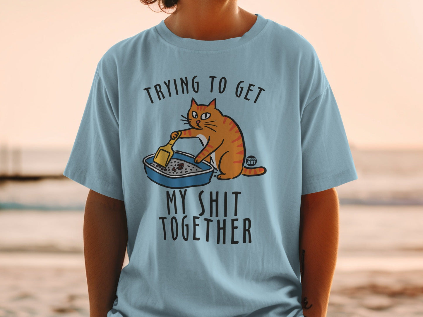 Light blue t-shirt with orange cat cleaning litter box, text "Trying to get my shit together".