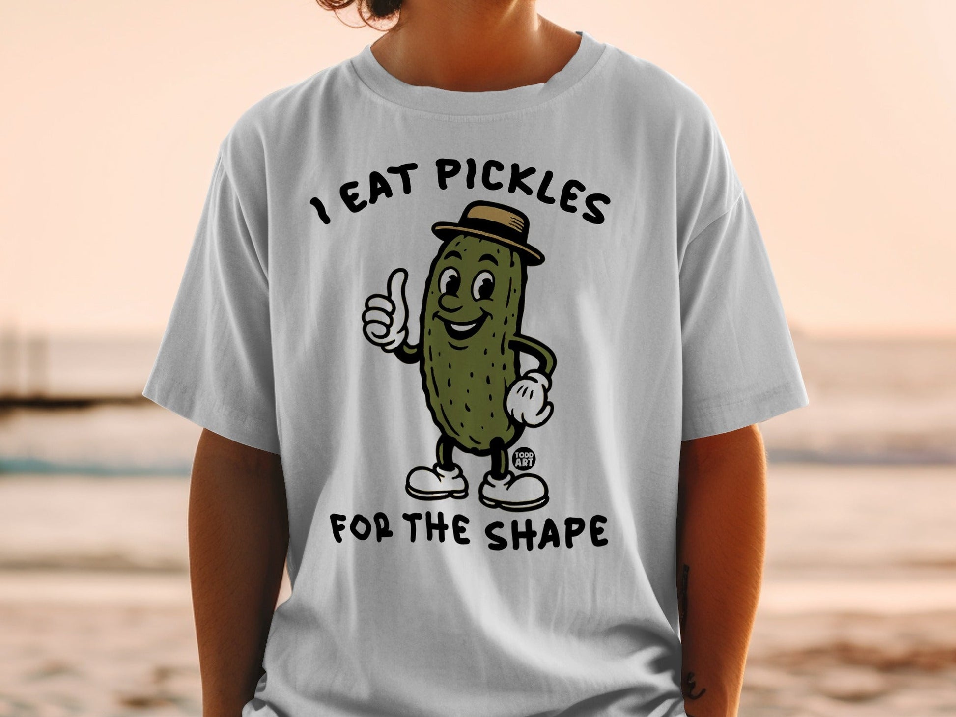 White t-shirt with cartoon pickle giving thumbs up, text "I EAT PICKLES FOR THE SHAPE".