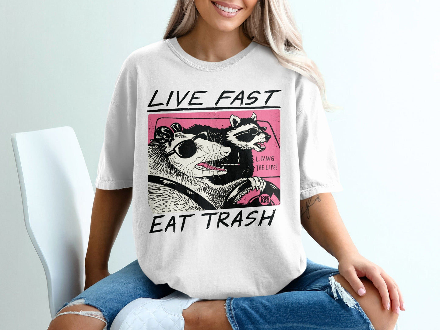 T-shirt graphic of possum and raccoon driving, "Live Fast Eat Trash" text.