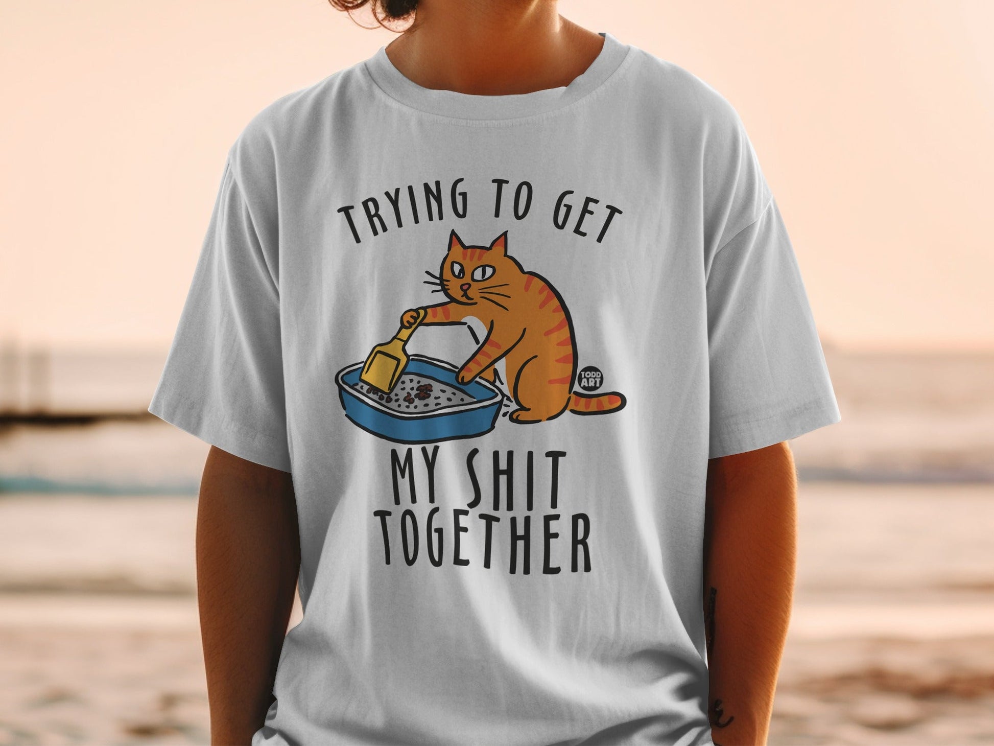 T-shirt with cartoon cat cleaning litter box and text "Trying to get my shit together".