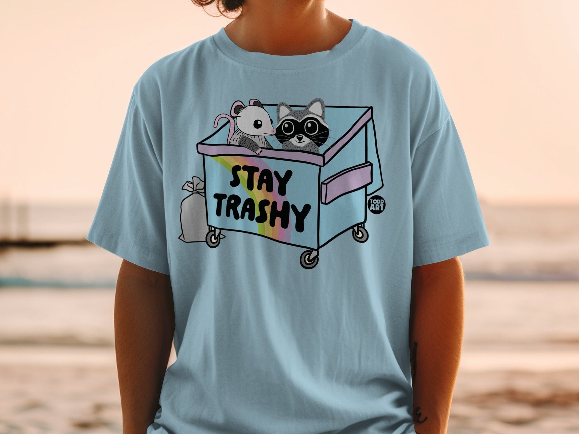 Light blue t-shirt with a cartoon raccoon and mouse in a trash can, "STAY TRASHY" text.