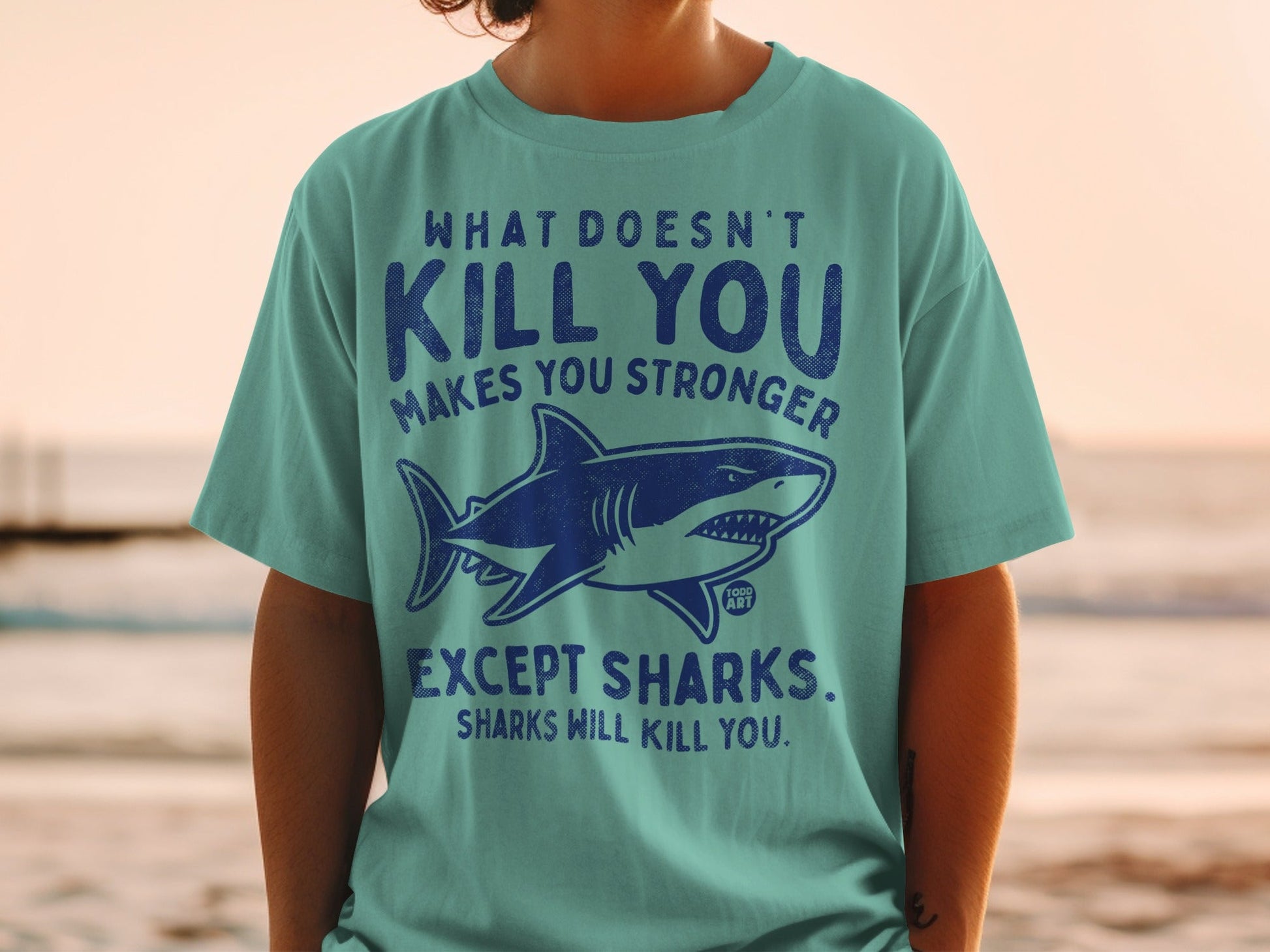Teal t-shirt with shark graphic and text "What doesn't kill you makes you stronger. Except sharks. Sharks will kill you."