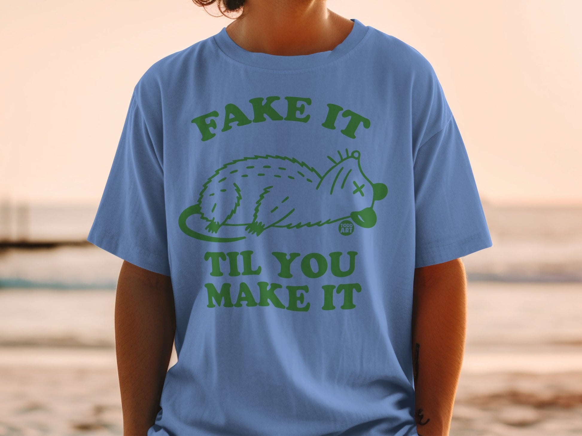 Blue t-shirt with green "Fake It Til You Make It" text and possum graphic.