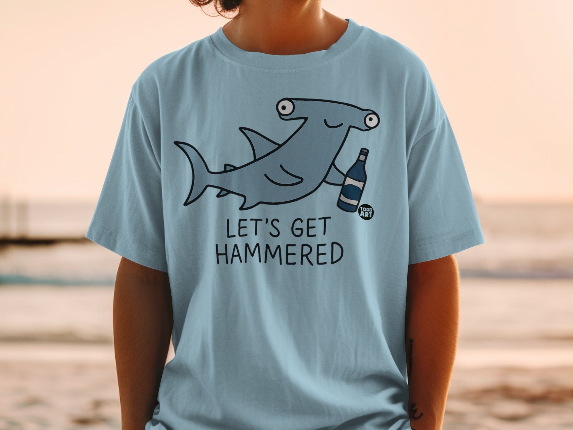 Light blue t-shirt with a cartoon hammerhead shark holding a bottle, text "LET'S GET HAMMERED".