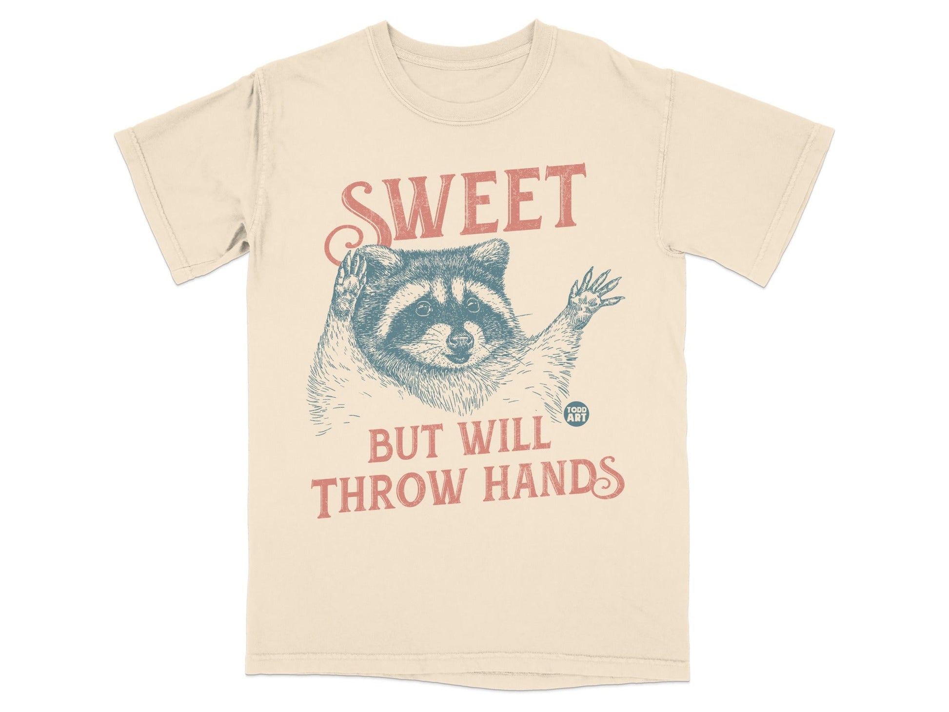 Cream t-shirt with raccoon illustration and text "SWEET BUT WILL THROW HANDS".