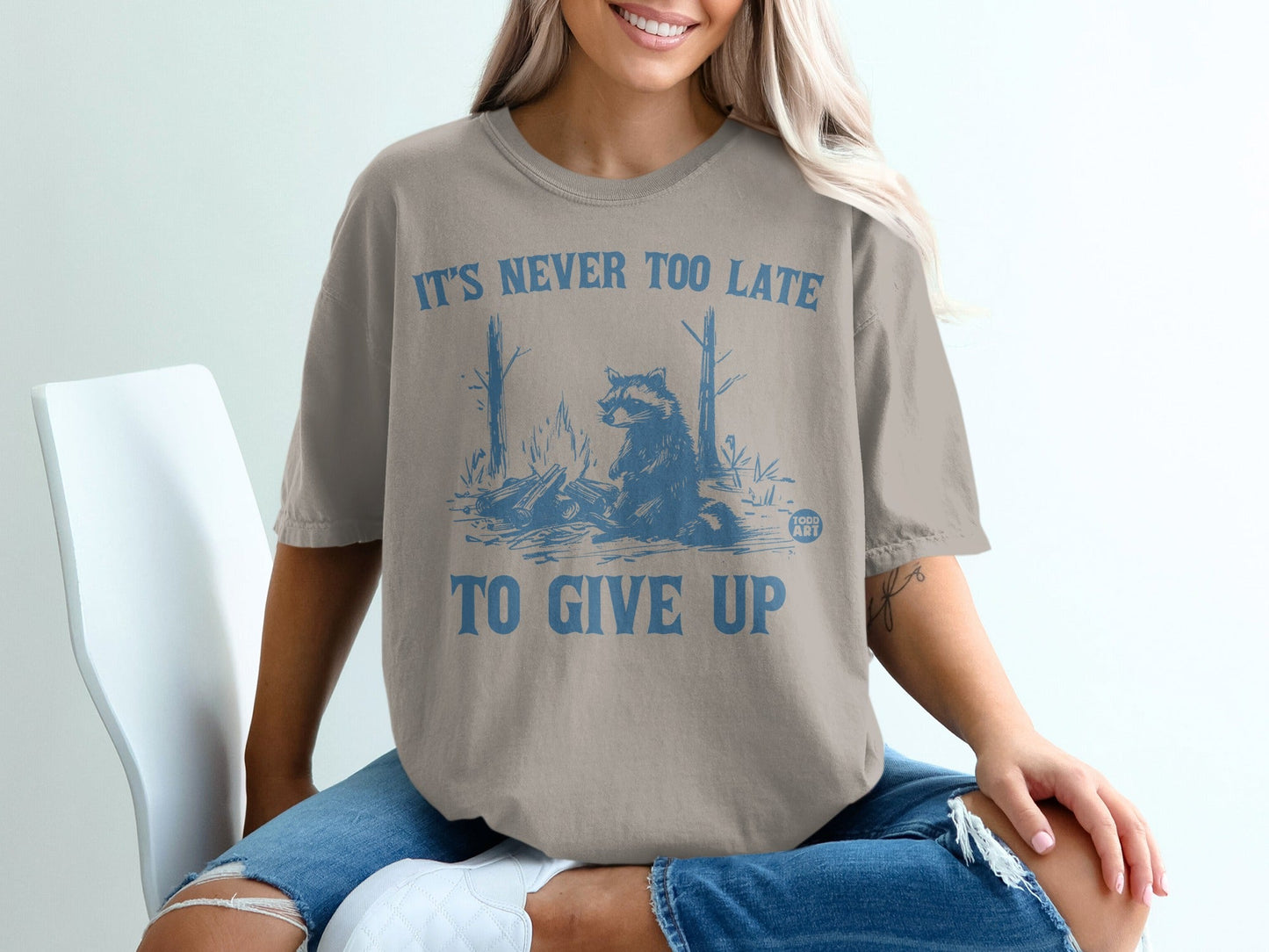 T-shirt with raccoon by campfire graphic and "IT'S NEVER TOO LATE TO GIVE UP" text.