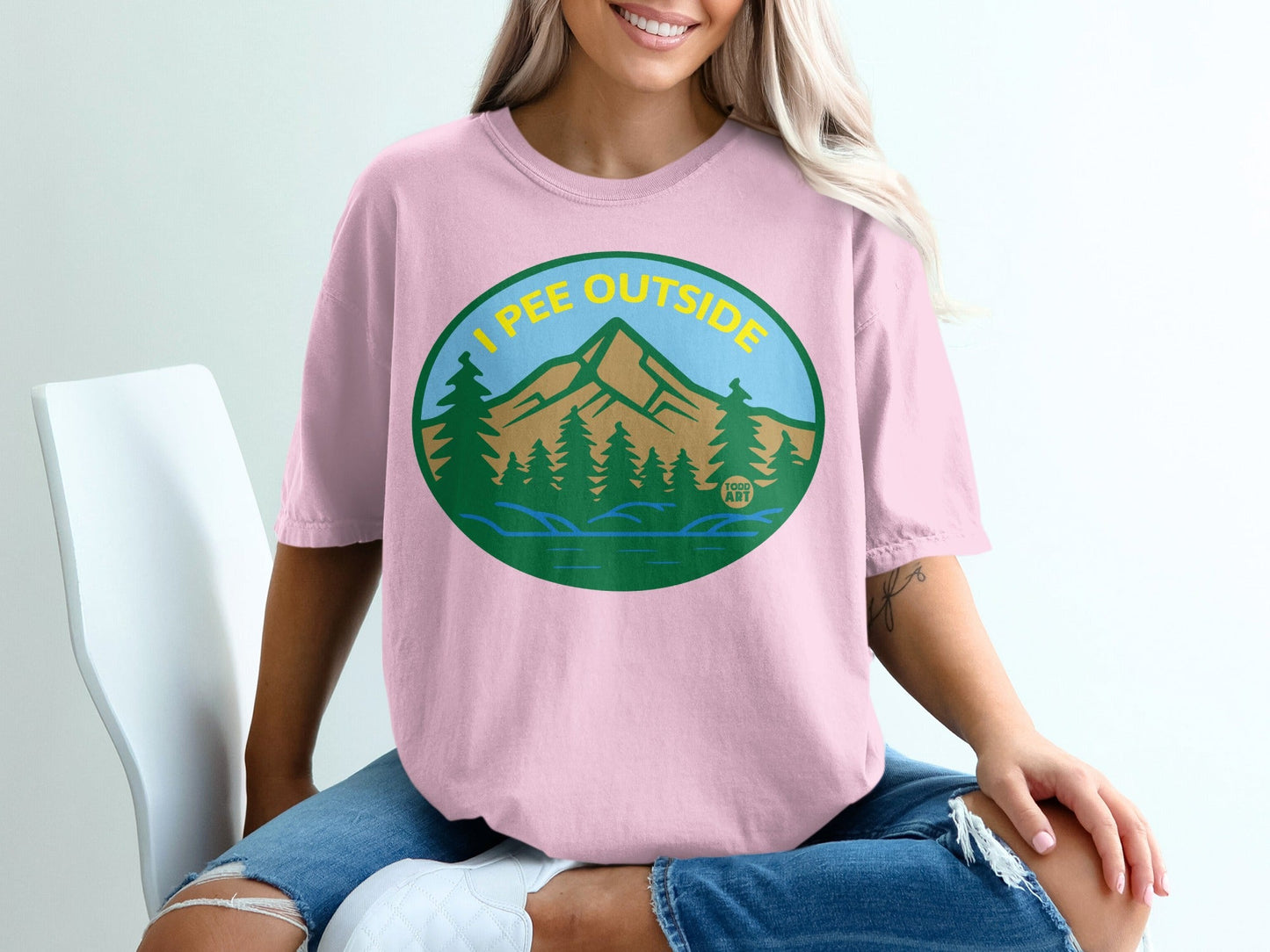 Pink t-shirt with oval design of mountains, trees, and lake; "I PEE OUTSIDE" text.