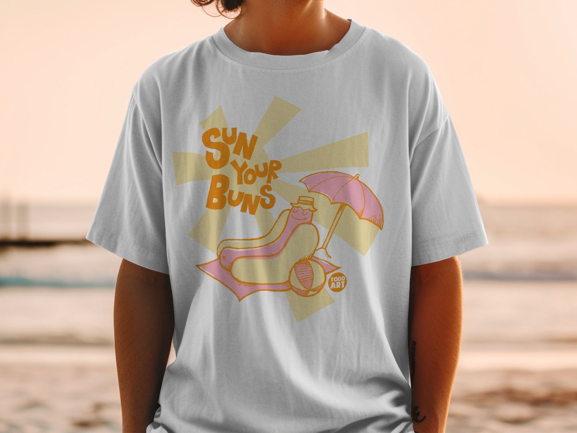 T-shirt featuring a hot dog with a pink umbrella on a beach towel.