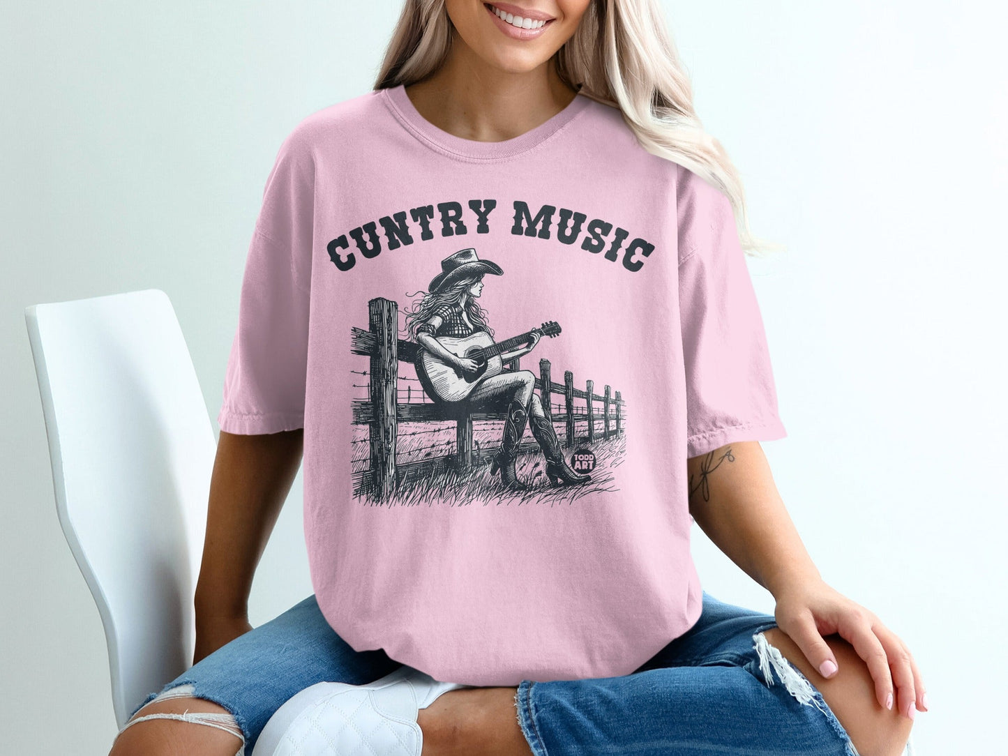 Pink t-shirt with black graphic of a woman playing guitar by a fence.