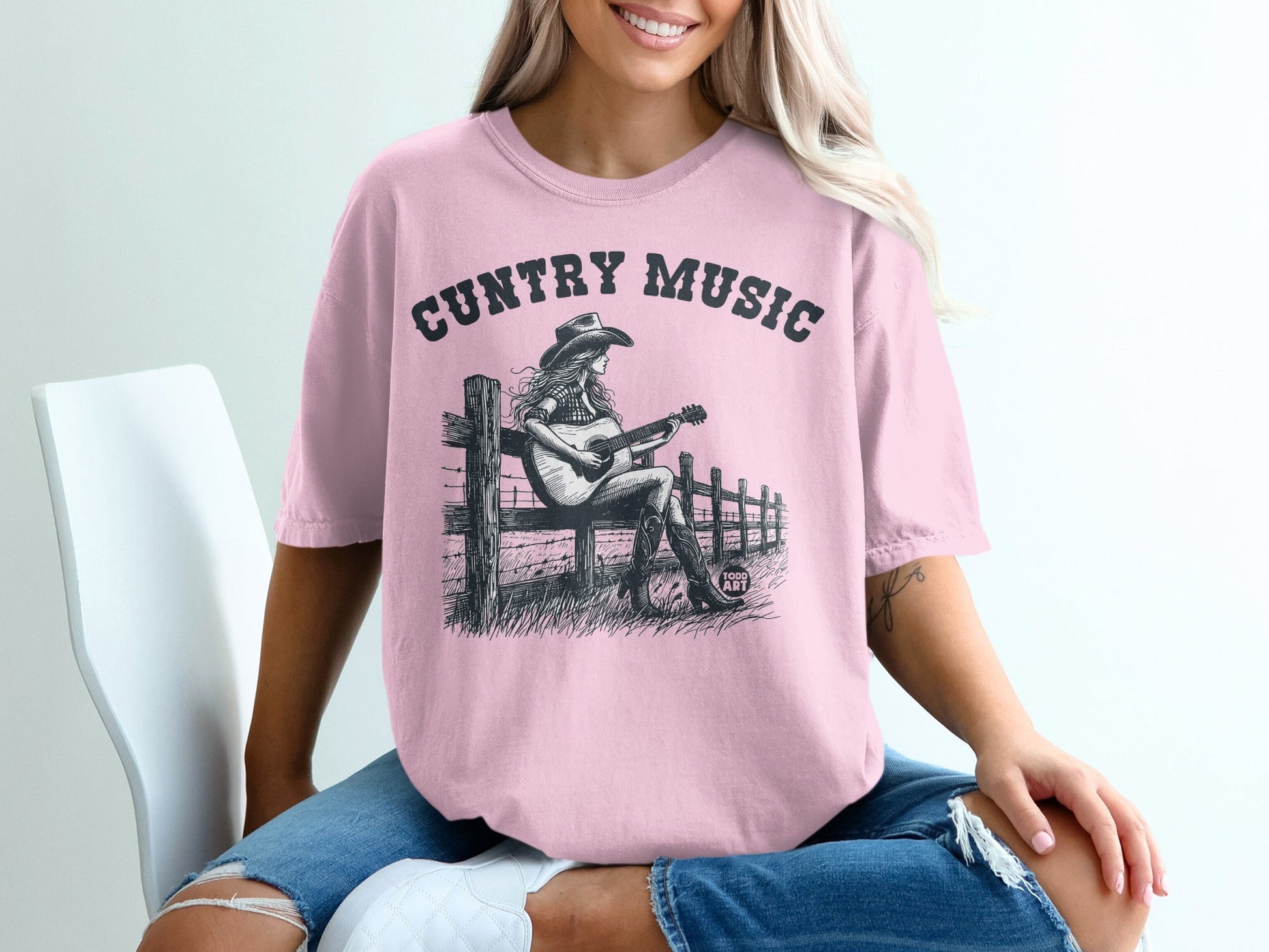 Pink t-shirt with black graphic of a woman playing guitar by a fence.
