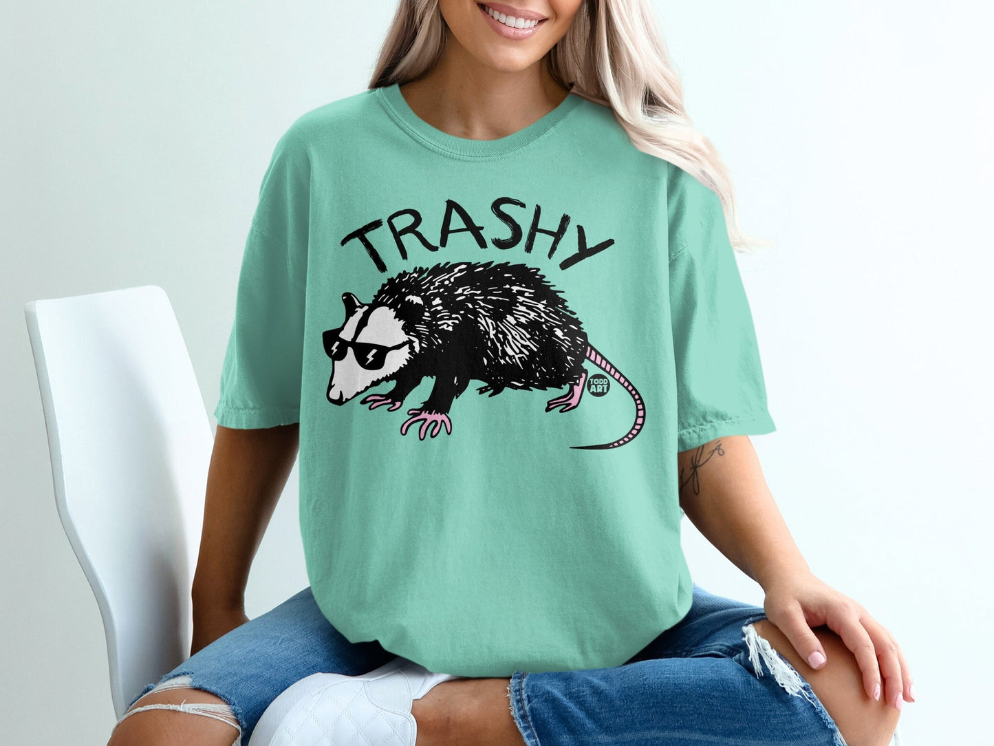 Opossum wearing sunglasses on teal t-shirt with "TRASHY" text.