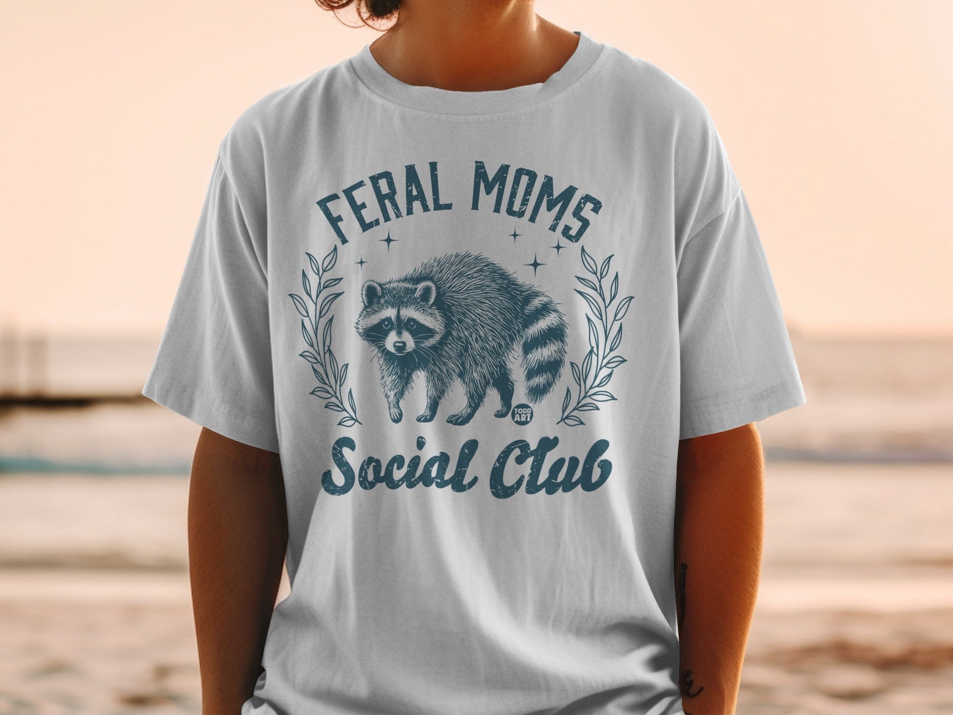 Feral Moms Social Club graphic with raccoon and laurel wreath on t-shirt.