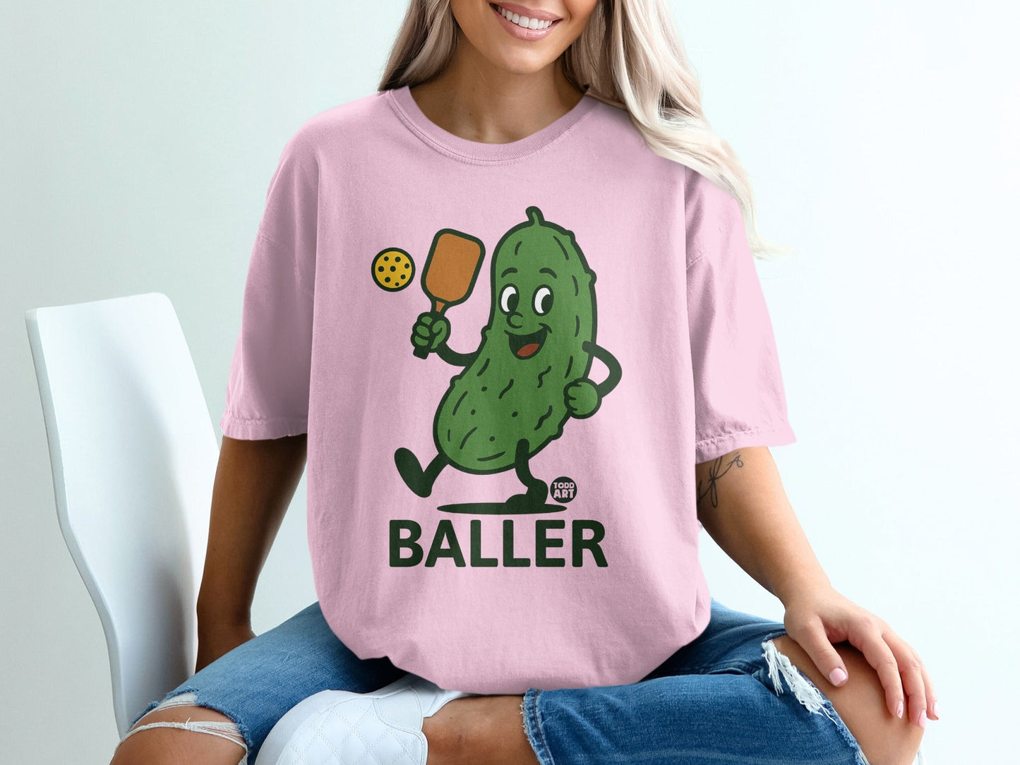 Pink t-shirt with cartoon pickle holding a pickleball paddle and ball.