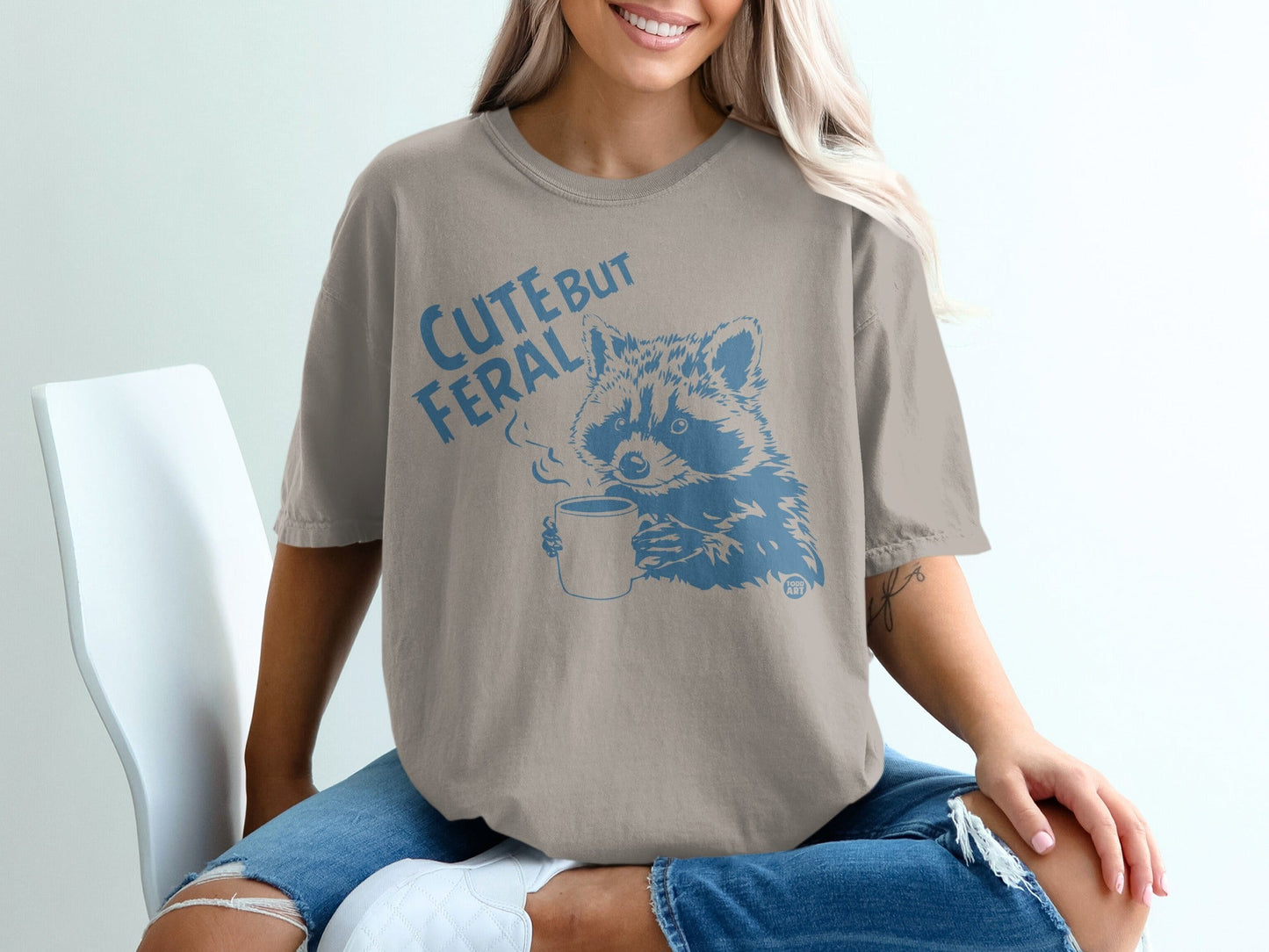 Raccoon with steam from mug, "Cute but Feral" text on taupe t-shirt.