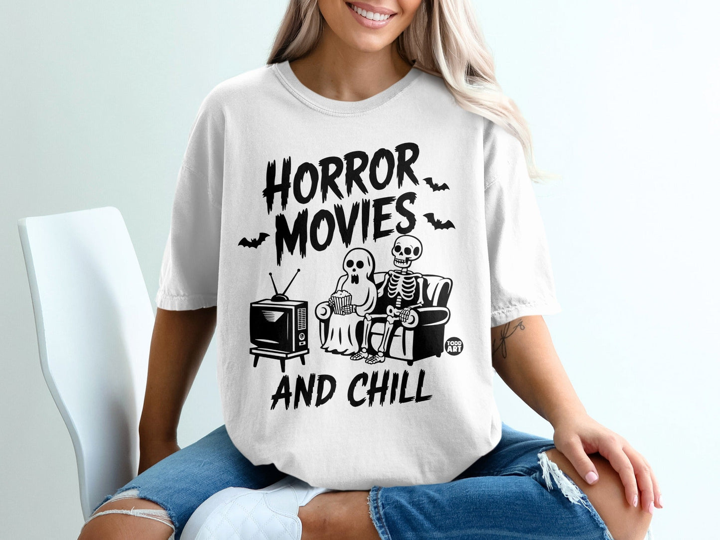 White t-shirt with "Horror Movies And Chill" text and cartoon skeletons watching TV.
