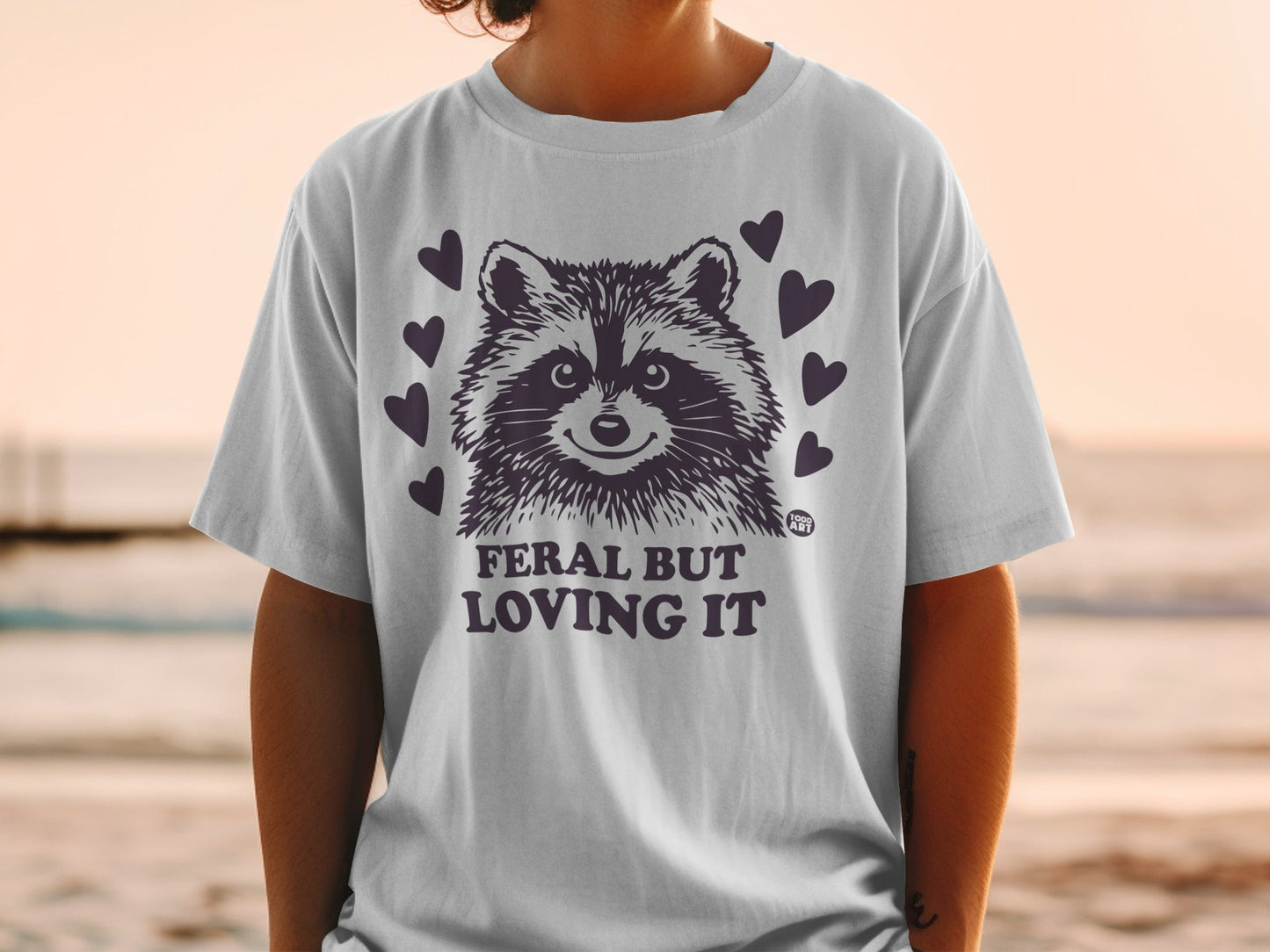 White t-shirt with raccoon graphic and text "Feral But Loving It".