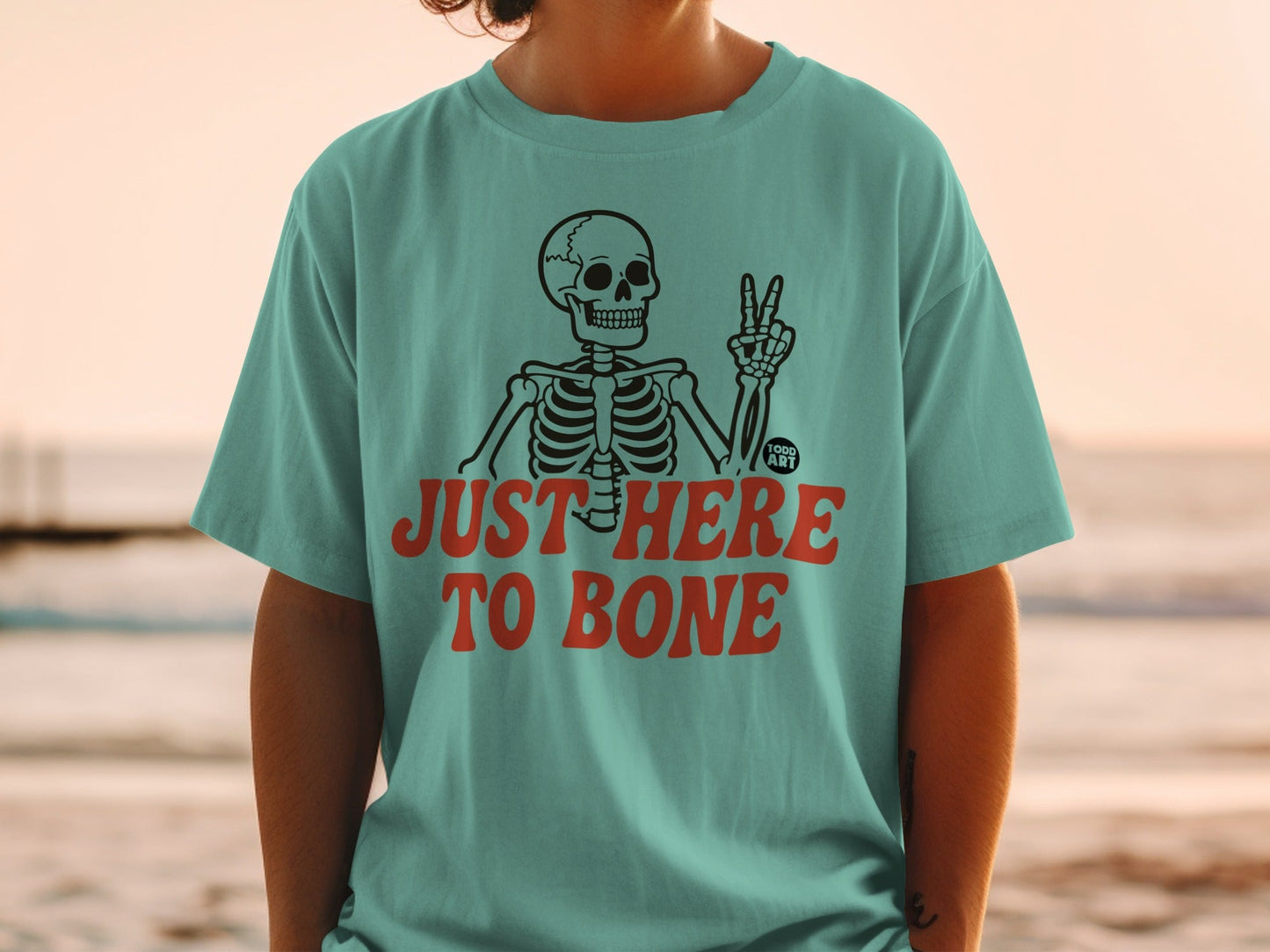 Teal t-shirt with skeleton giving peace sign and text "JUST HERE TO BONE".